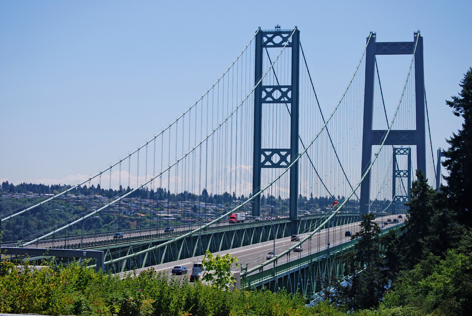 Twin suspension bridges, known as the Tacoma Narrows Bridge, on a sunny day. Greenery is seen in the foreground, and cars drive over each bridge in the correct direction.