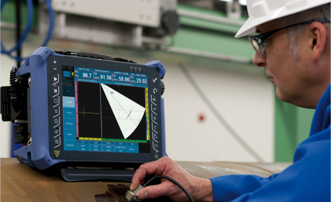 Technician wearing a white hard hat and blue shirt, and glasses, uses Phased Array UltraSound to examine a weld for defects. The technician holds a device over a weld. On a screen in front of the technician a projection of the ultrasound appears.