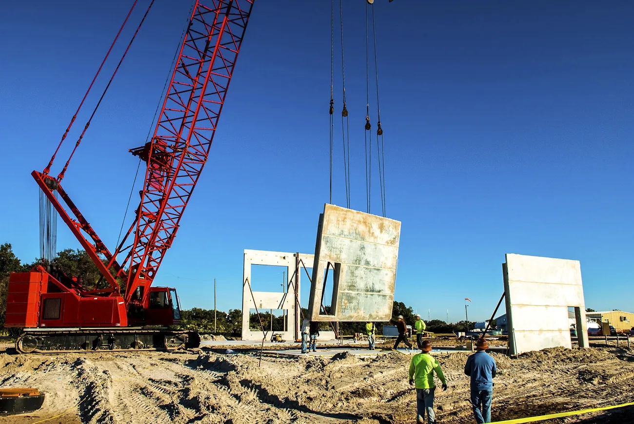 A red lattice boom crane mounted on crawler treads lifts precast tilt-up concrete panels into place on a construction site.