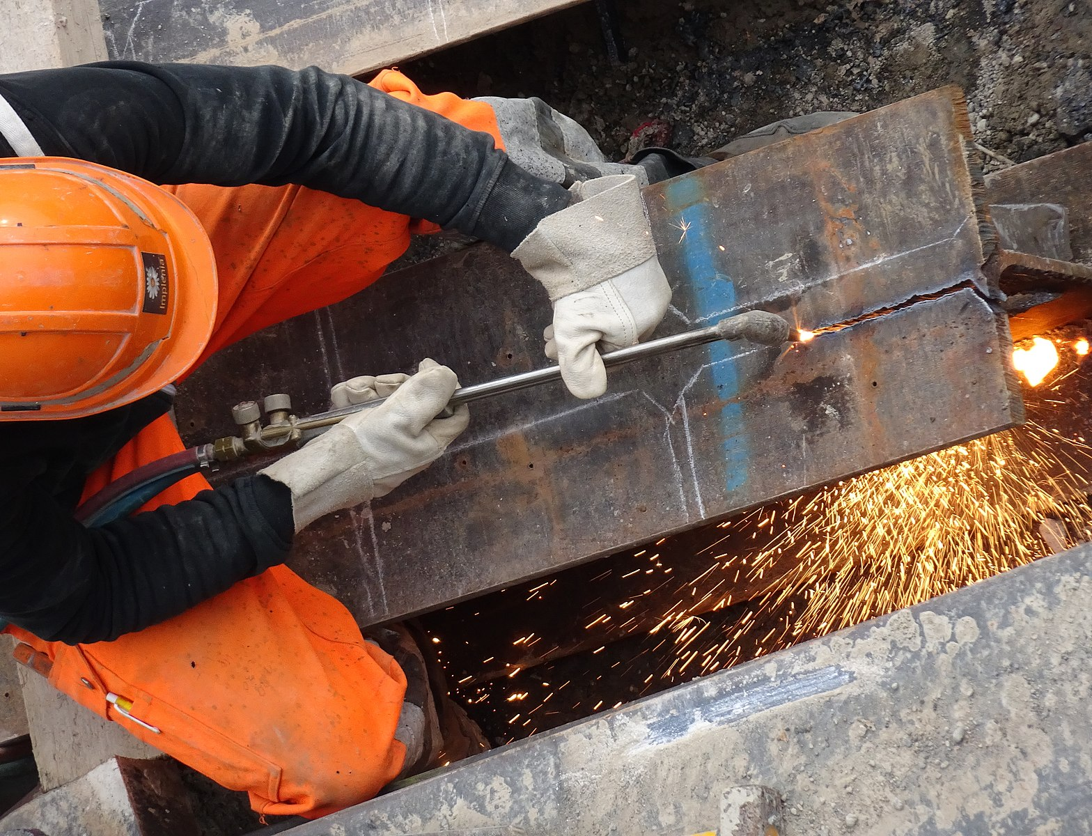 An overhead view of a worker wearing an orange helmet, orange protective pants, a black protective shirt, and gloves uses an oxy-fuel cutting setup to cut a shape out of a piece of metal.