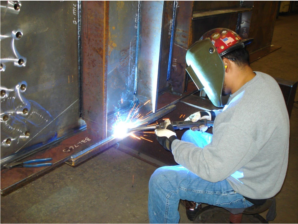 Welder wearing PPE, sitting on a stool while welding a steel girder