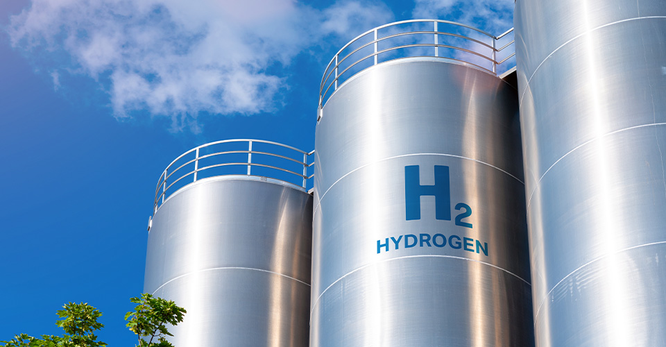 Stainless steel storage tank that looks like three shiny silver cylinders with guard rails on top. The center cylinder is labeled in big blue letters H2 Hydrogen.