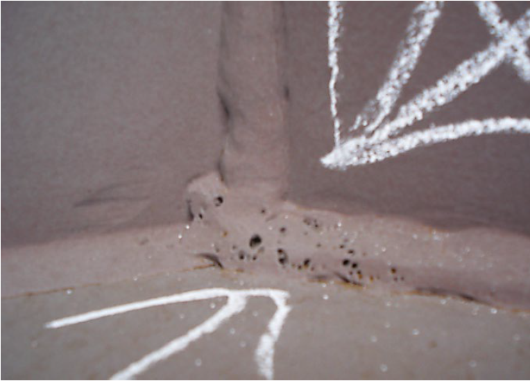 Porosity welding defect shown on a weld bead joining plates together. The weld bead has many small holes in it. Two white arrows drawn in chalk on the image point to the holes in the weld bead.