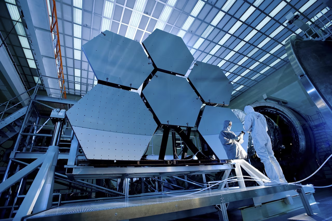 A worker wearing a white safety suit stands in front of the James Webb Telescope in a building. This view of the telescope includes six hexagonal shaped panels that are reflective. The worker is standing in front of one of the hexagons, so even though the camera is facing their back, we can see their front as a reflection in the hexagon.