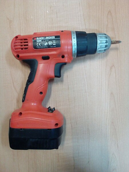 An orange, hand-held, battery-powered drill. The drill motor sits atop a handle at 90 degrees to the handle. A rectangular battery pack is attached to the bottom of the handle. The handle has a trigger to start the drill. At the working end of the tool, a driver bit is installed meant to drive screws.