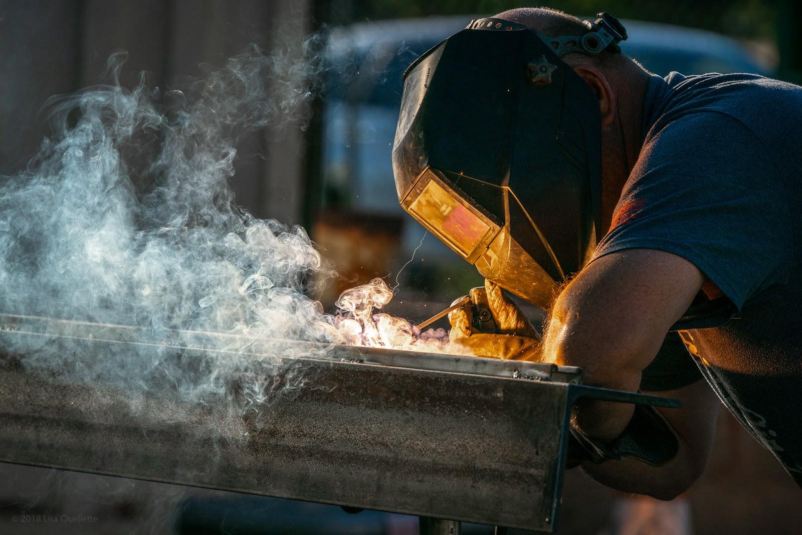 A man performs SMAW welding in a T-shirt, gloves, and a welding hood.