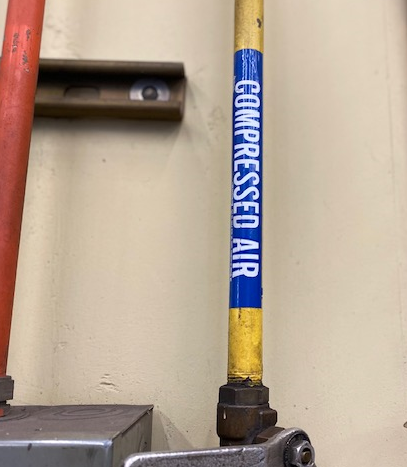 A yellow pipe runs along a shop wall. A blue label on the pipe, with white lettering, reads compressed air. Piped in compressed air aka ‘shop air’ is used in CAC-A to blow away dross and molten metal.