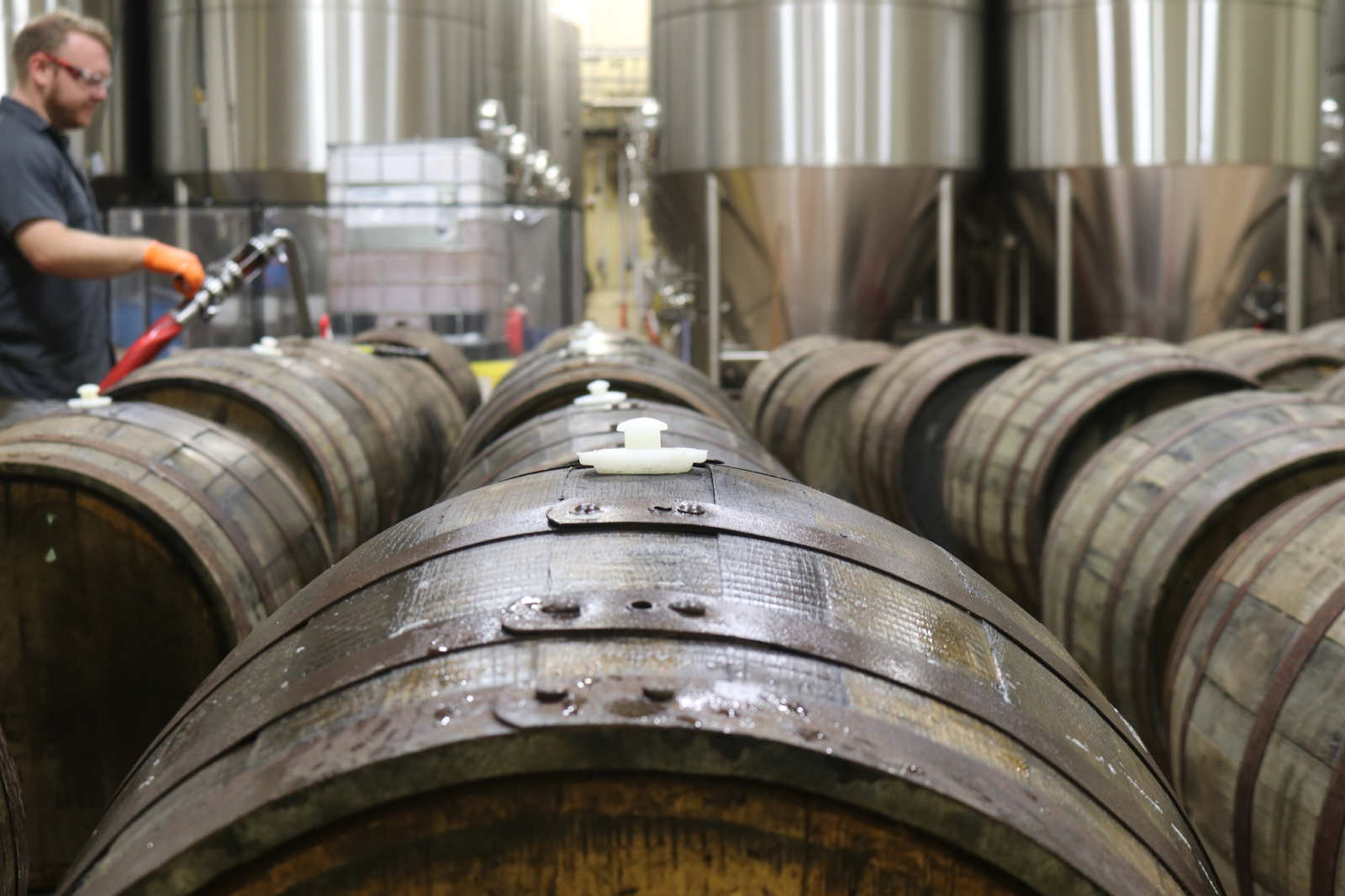 Behind a series of wooden casks used to age beer is a collection of stainless steel tanks, which are taller than everything around them. To the left a worker handles an instrument used in beer making.