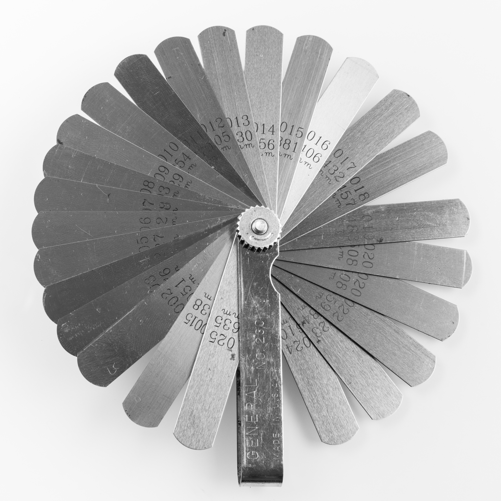 Photograph of a set of feeler gauges fanned out. Each feeler gauge is a thin strip of metal with a measurement printed on it. The gauges are held together with a nut at the base so that each gauge can be inserted in a gap but stay attached to the entire set.