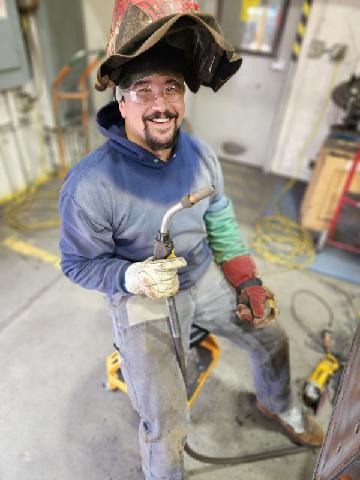 A welder holding a torch with the hood of his welding helmet tipped up sits on a stool and smiles.