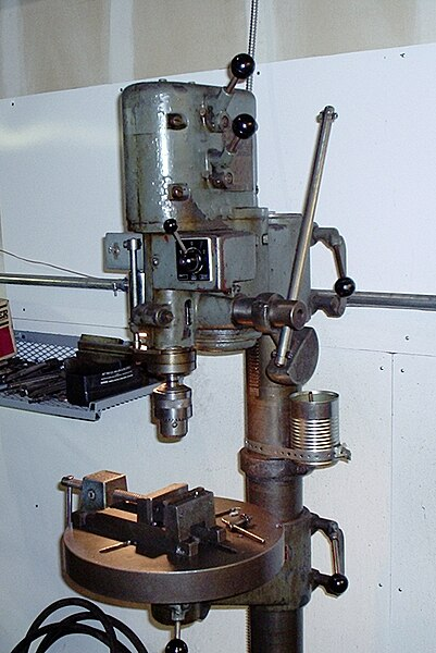 A large upright drill press made out of metal. The drill motor sits atop a stand with the spindle pointing downward. There is a round table with a vice attached to the stand underneath the drill, where the material being drilled can be affixed and clamped. There is a chuck attached to the spindle for holding different sizes of drill bits, and a lever for lowering the bit into the material being drilled.