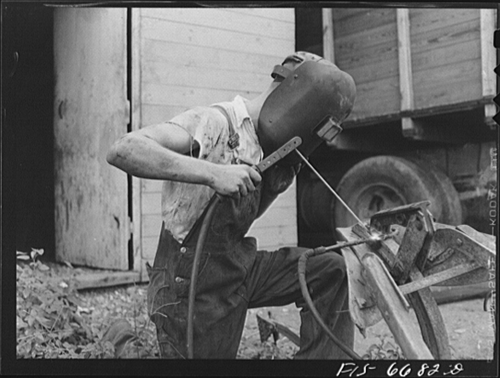 A farmer, wearing short sleeves, denim overalls, and welding mask leans over a metal part of a tractor or trailer as he welds a part, presumably doing a repair. He is welding outdoors.