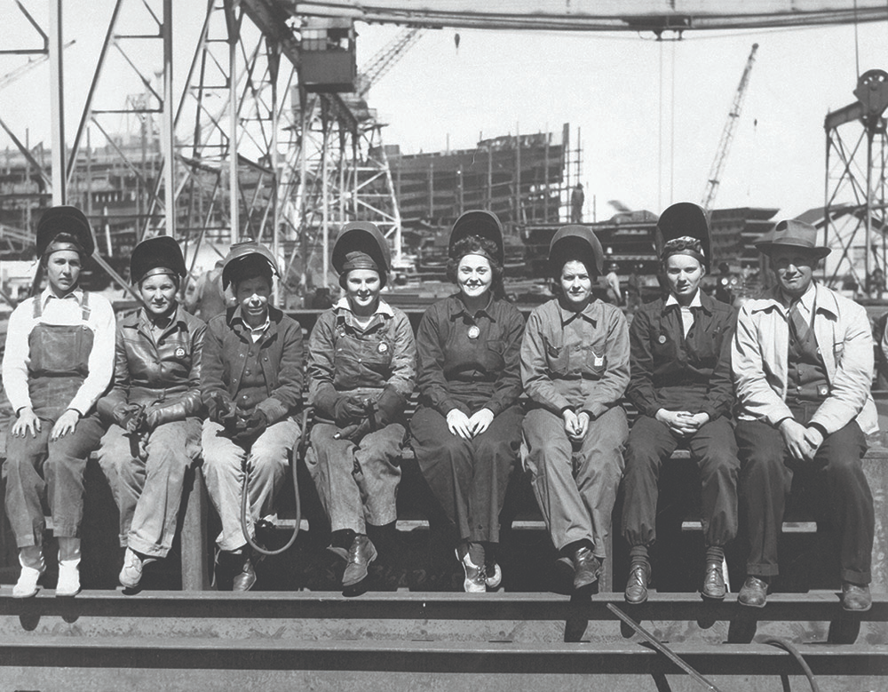 Seven women wearing welding helmets, long sleeves, long pants, and boots with one man sitting on the far right sit in a line and pose for a picture in a ship yard.