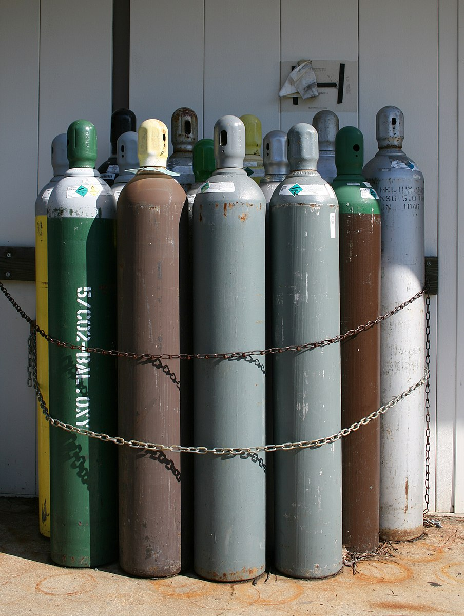 Several high-pressure gas cylinders of different colors are stored against a wall, secured by a chain around them.