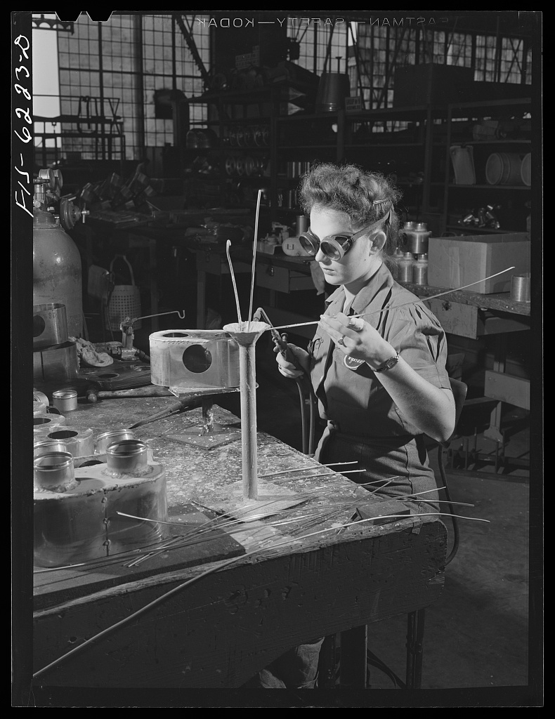 A woman wearing safety glasses sits at a worktable in a shop. She holds a torch in her right hand and a wire in her left hand as she welds a part for a fuel pump.