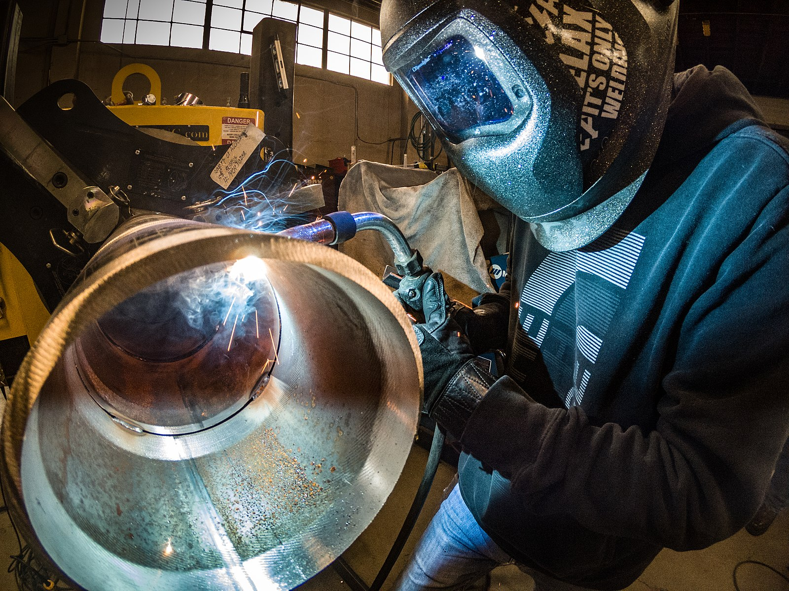 A welder in a black sweatshirt and helmet uses Gas metal arc welding on a pipe in the 5G position