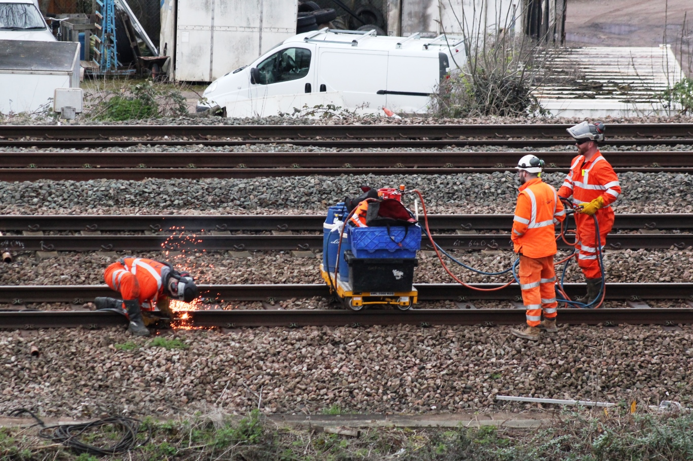 On the left of the photograph, one worker uses an OFC process to cut rail with two safety observers. Between the worker cutting and the safety observers is a portable cart with a power source and fuel tanks for cutting. The safety observers hold one blo\ue and one red hose away from the person welding.