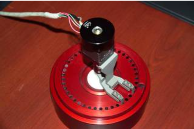 Eddy Current Probe sitting on a desk. The probe has a large red base with a smaller black cylinder on top of that. There is a cord leading out of the black cylinder.