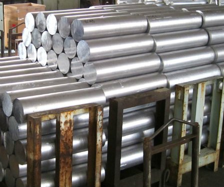 Aluminum bar stock is piled on three metal holders made to hold the stock in place. There are hundreds of pieces of aluminum. Each piece is at least three feet long and in the form of a cylinder with a radius of about 1 foot.