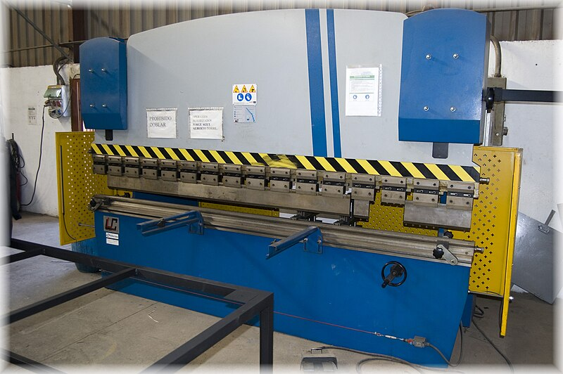 A blue, gray, and yellow machine used to bend sheet metal. The machine is rectangular, about six feet tall and ten feet wide, with a horizontal opening in the middle. The top part of the machine is made to move toward the bottom part like a set of jaws. The space between the top and bottom is where preshaped dies can be placed to bend metal to a specific angle or radius. The yellow parts of the machine are guards which are set to the sides to keep body parts away from the jaws when in operation.