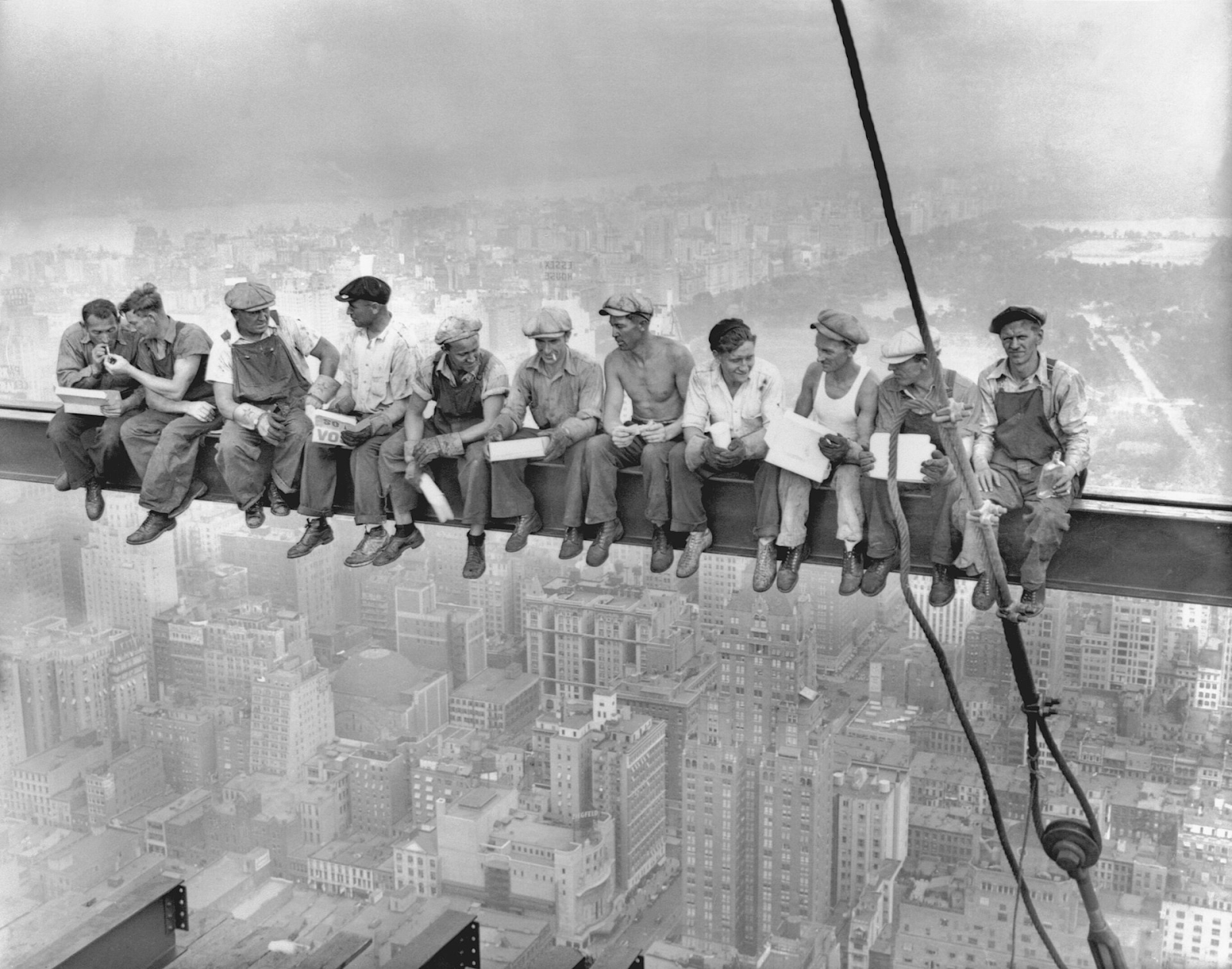 Several construction workers are seated on a beam high atop a skyscraper, chatting while eating lunch.