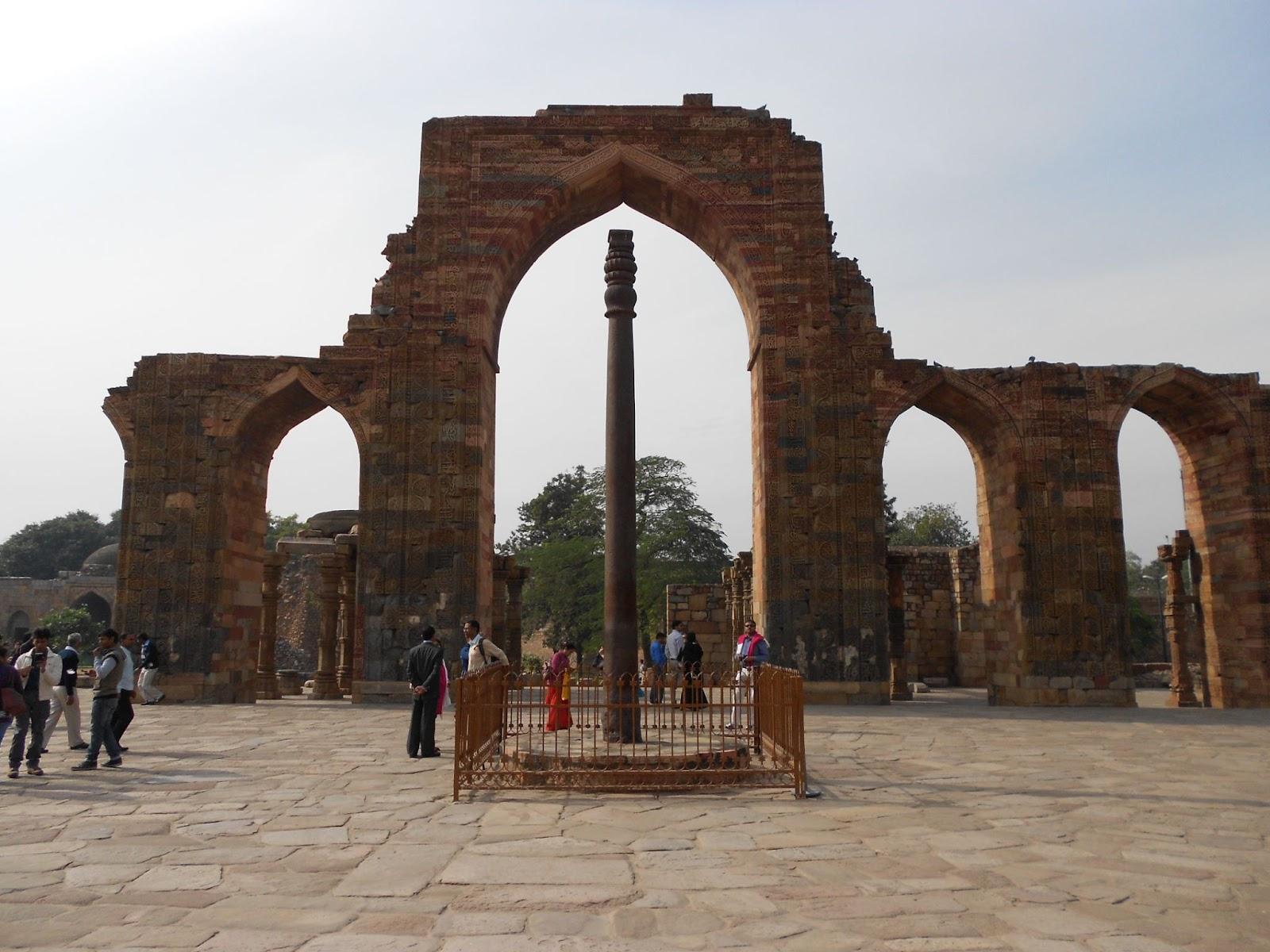 A cylindrical column is being admired by tourists, with ancient ruins in the background.