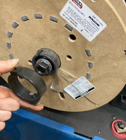 A side view of the wire reel attached to the machine, it is secured by a plastic clip that holds the reel in place when the machine is working. A hand demonstrates how to release the clip by squeezing the two metal buttons on the sides of the clip.