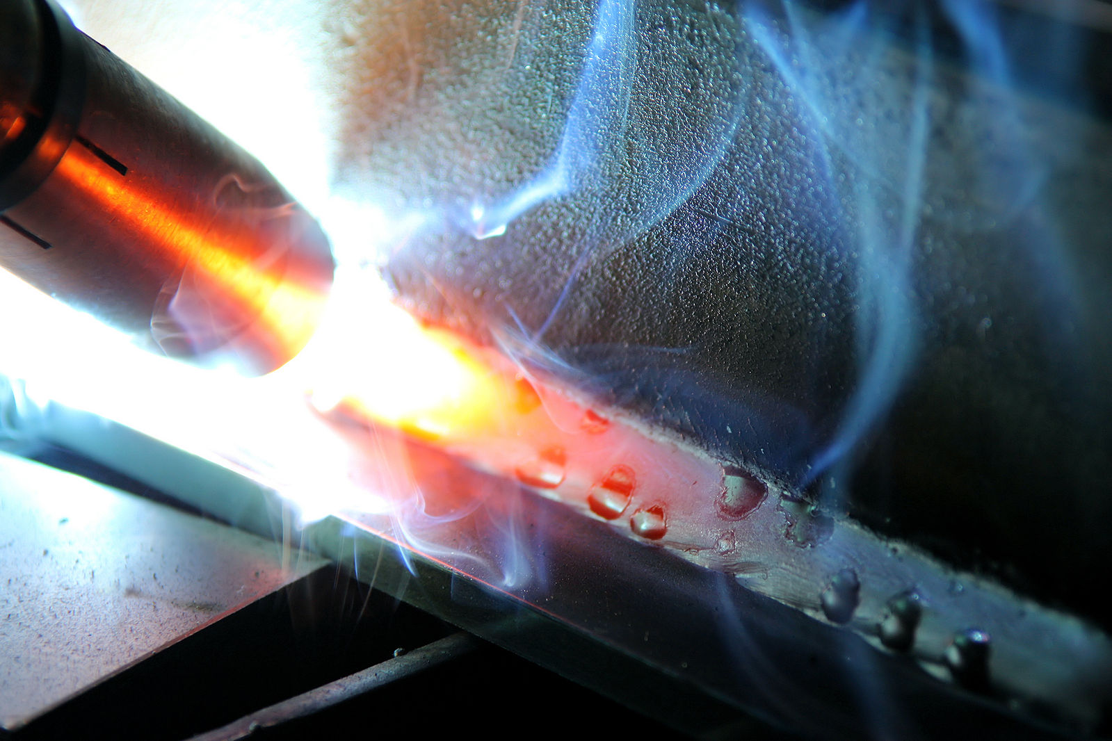 A weld being created in the corner of two intersecting plates, one plate lies flat on the table, while the other metal plate is perpendicular, forming a ‘T-joint’. The images shows the copper GMAW nozzle in the upper left corner with the weld trailing behind in a ‘pull’ travel direction using the GMAW spray transfer mode.