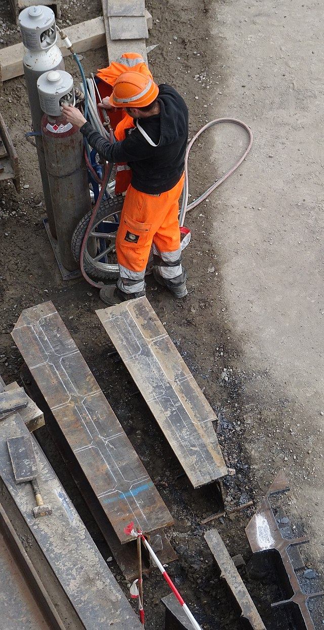 A worker wearing safety gear is setting up a workspace by checking gauges on gas canisters before making cuts.