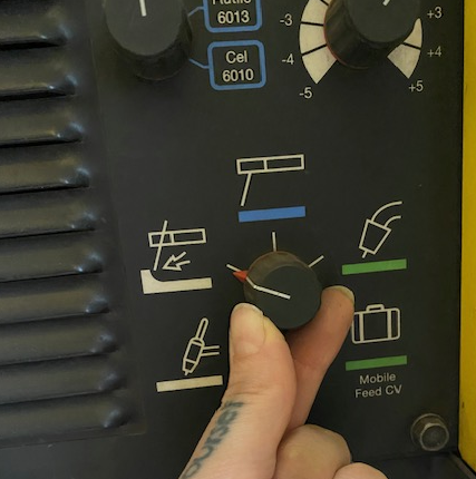 A dial on a welding machine showing different settings for the machine. A welder is moving the dial to the CAC-A setting. The picture of the CAC-A setting on this panel is a rectangle (resembling two pieces of rectangular metal that were welded together, with a line bisecting it representing a cut into the metal.