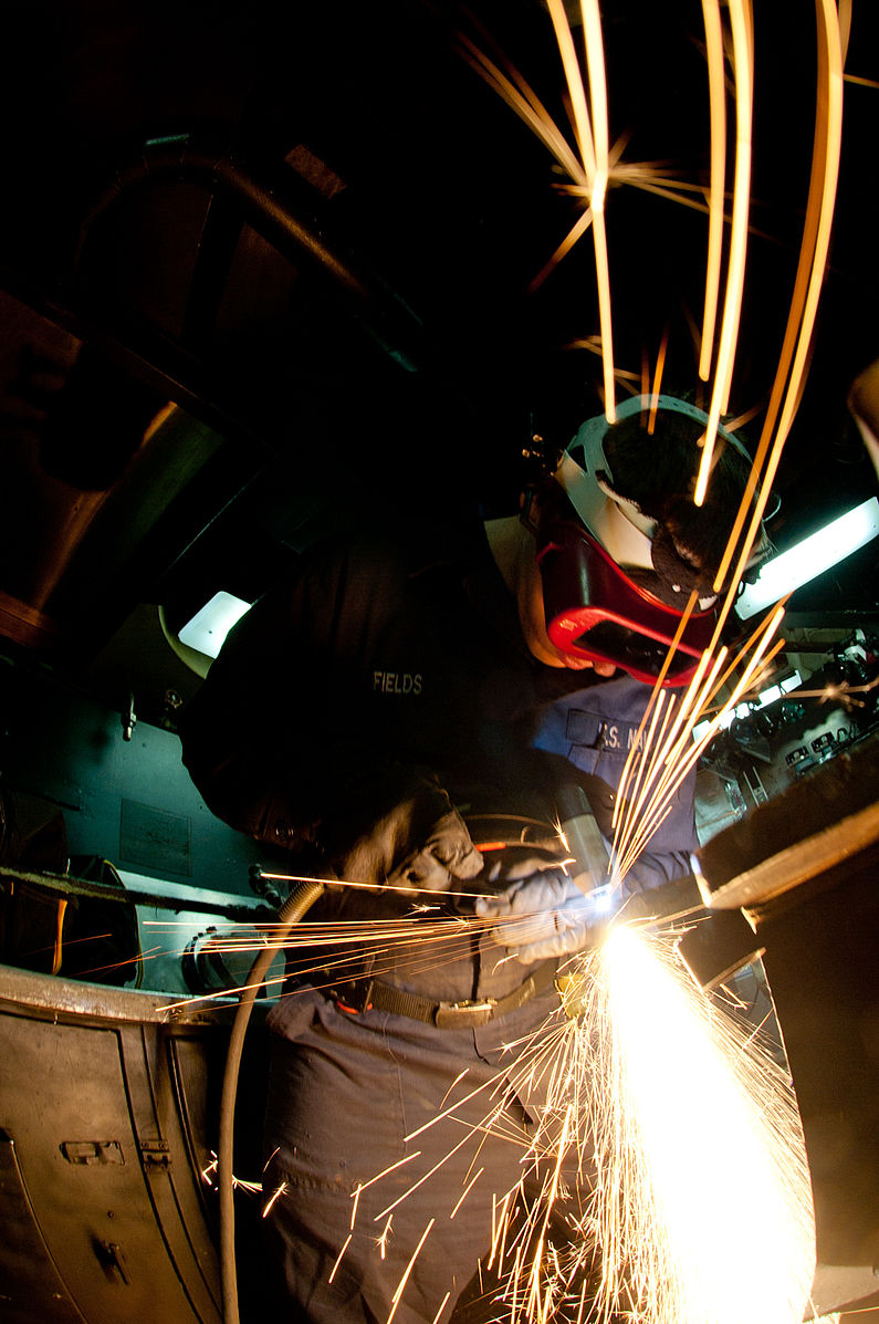 a welder wearing a protective lens, leather gloves, a long sleeved shirt, and long pants, using PAC to cut into a piece of metal. There are sparks from plasma cutting material in the image.