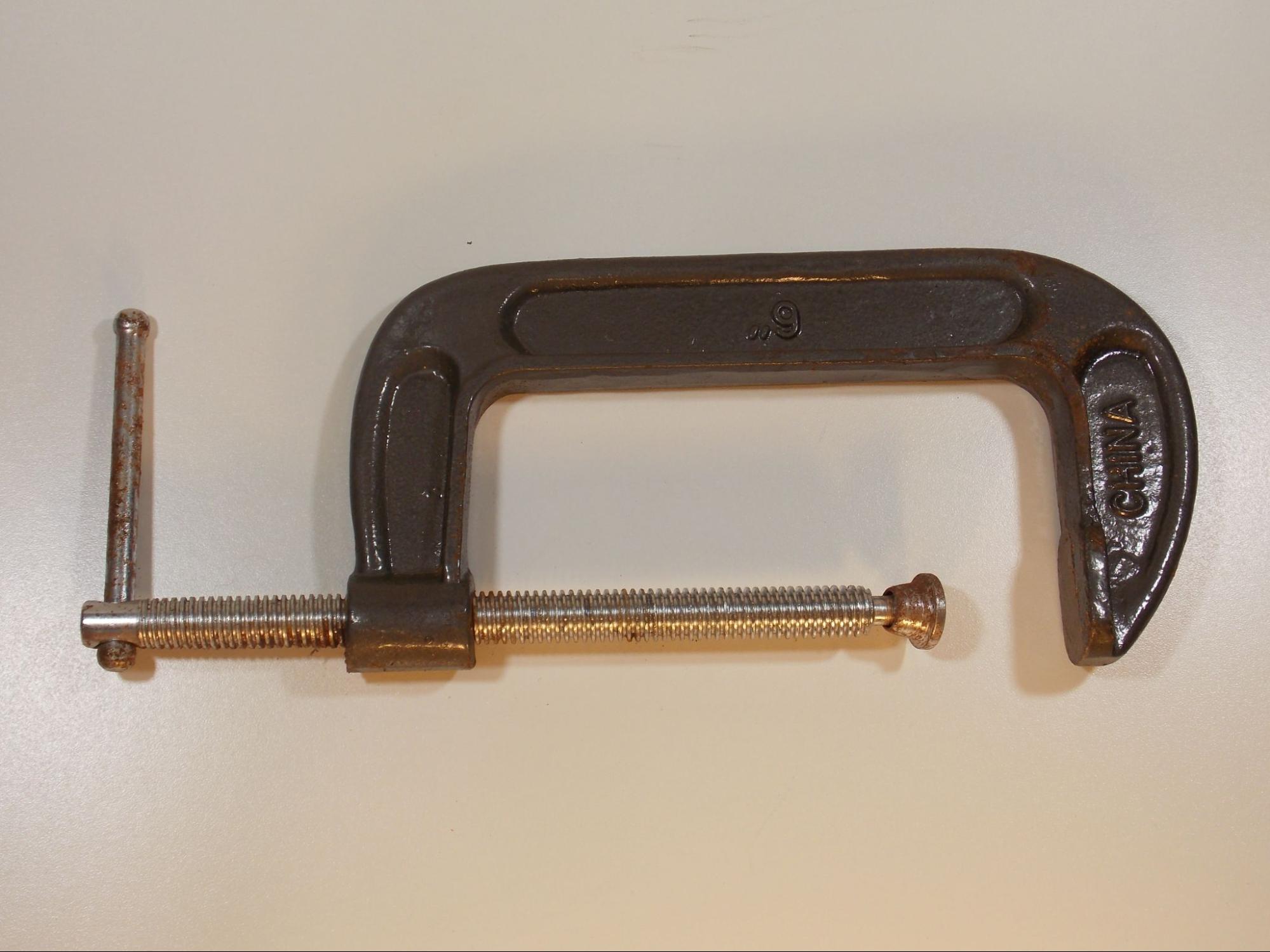 A 6 inch C-clamp. The main part of the tool is shaped like the letter C, with the top end of the C flattened so it can hold the material being clamped. The bottom end of the C has a threaded hole in which a large threaded screw is inserted. At the top end of the screw is a small flat foot that compresses the parts being clamped against the flat part on the C. At the base of the threaded screw is a small metal bar which is perpendicular to the screw. This is used for leverage so that the screw can be tightened until the clamp tightly compresses the material being held.