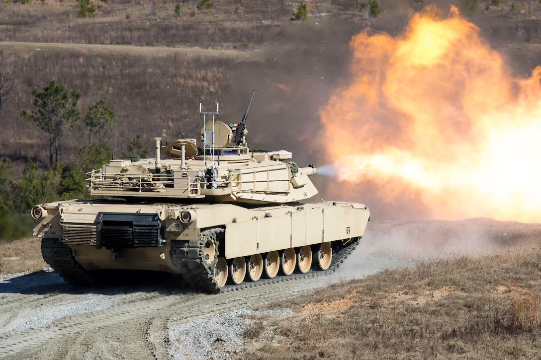 A sand-colored military tank with treads, armor plating, and a gun mounted on the top. An explosion of fire and smoke proceeds the tank, showing that it has fired the canon. The tank is in a track with sparse vegetation surrounding it.