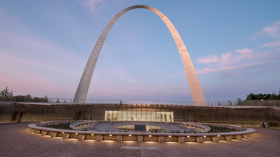 St. Louis Gateway Arch is pictured rising over the visitors’ center and museum building that is underneath the arch. Behind the arch the sky is blue with few clouds.