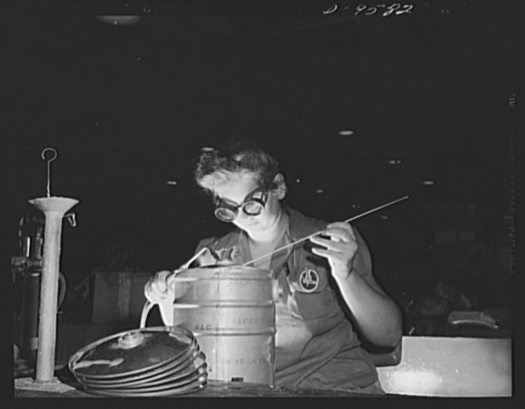 A woman from the World War Two era, wearing a pair of protective glasses, and sitting comfortably at a work table holds a torch in her right hand and an electrode in her left hand as she focuses on welding an aluminum canister. This is an example of aluminum oxy-acetylene welding.