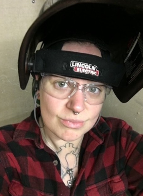 A women wearing safety glasses, a red and black flannel shirt with a welding helmet flipped up on her head