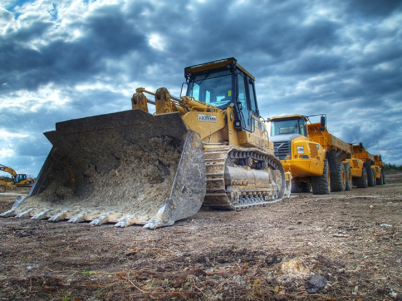 A bulldozer with a large bucket on the front is parked in front of two dump trucks.