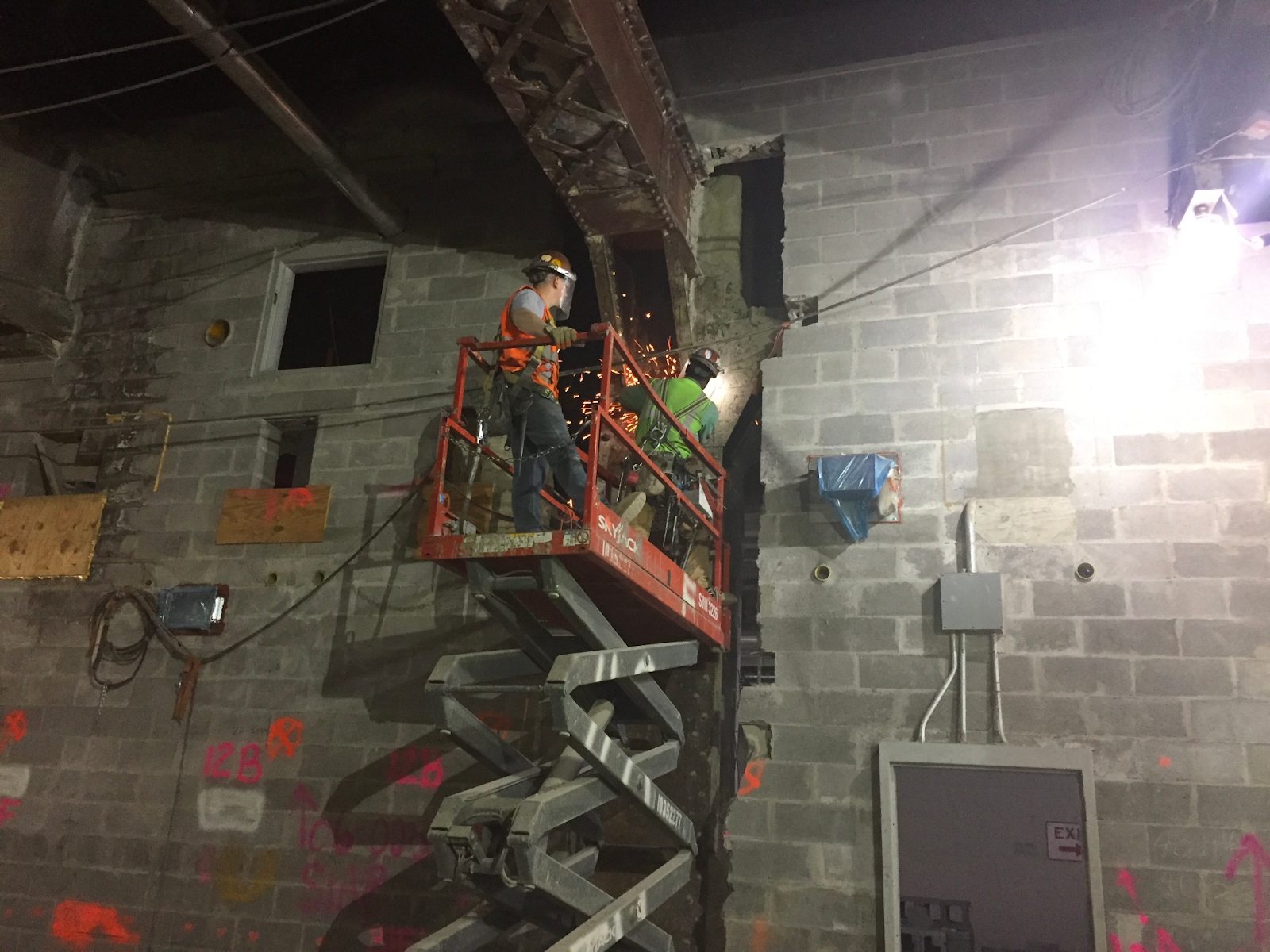 Two construction workers stand in a scissor lift that is raised off the ground. One of the workers is performing hot work on a girder, and sparks fill the air.