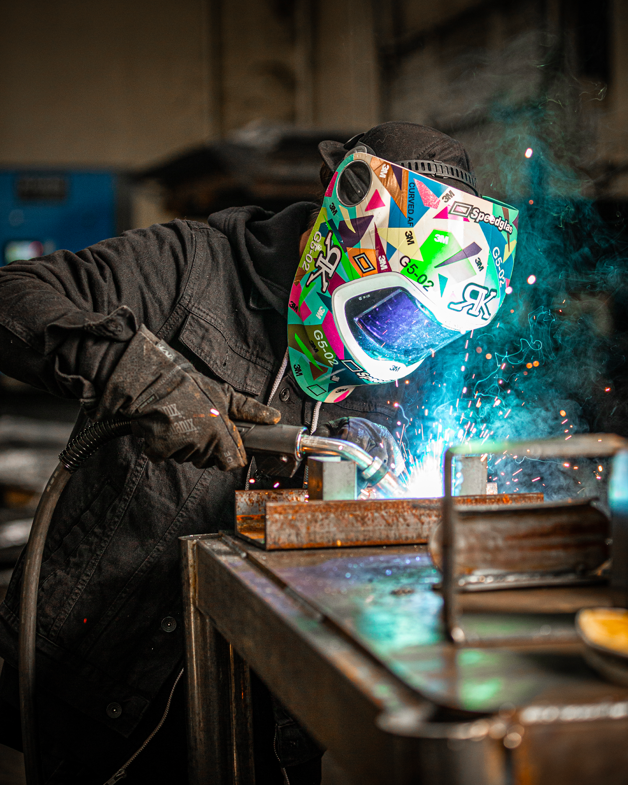 A welder in black coveralls and a colorful welding helmet uses gas metal arc welding to join together multiple pieces of metal in a shop.