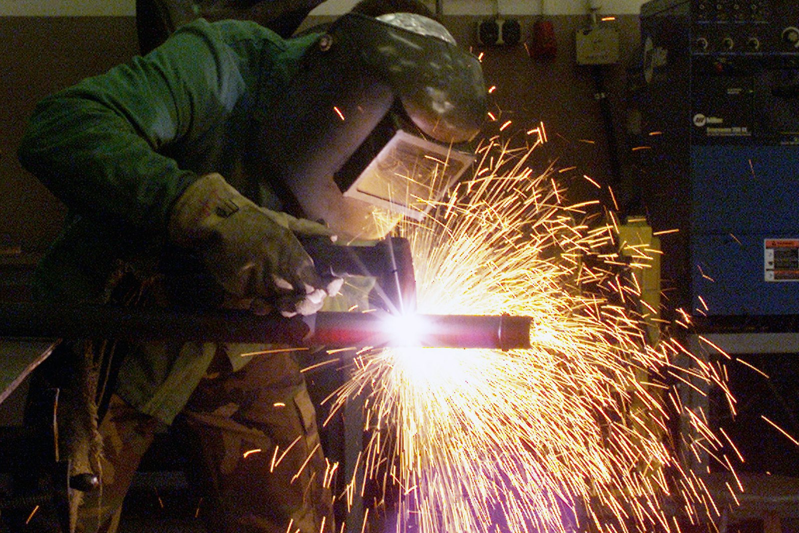 A welder wearing a welding hood with a lens, gloves, a long sleeved shirt, and protective pants, uses PAC to cut through a pipe-shaped metal object. Sparks surround the pipe and torch as the welder operates the torch.