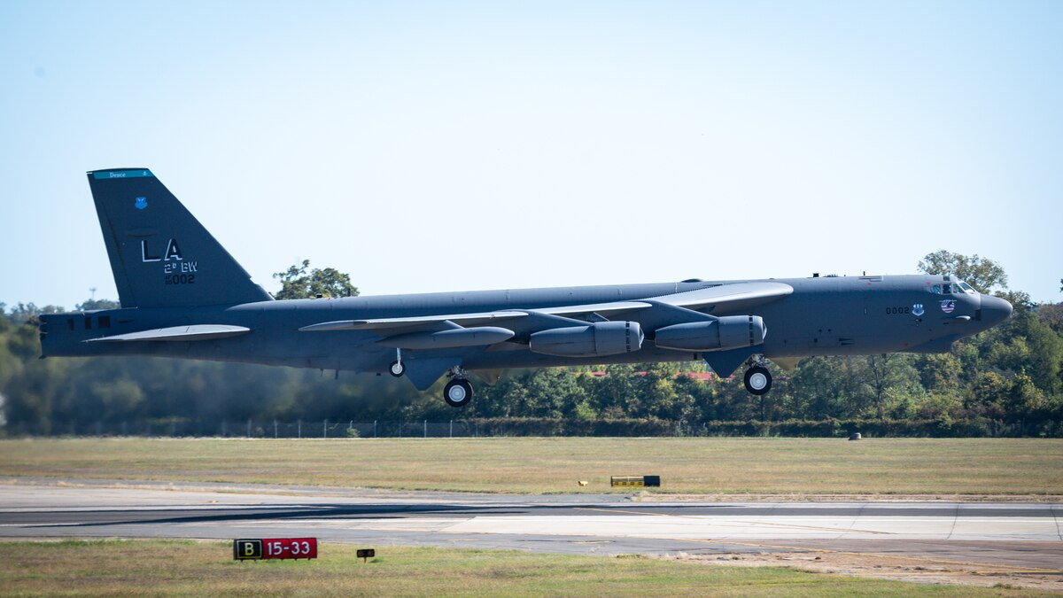 A grey-colored military jet with the letters LA printed on the tail of the jet taking off over a runway. The landing gear is still extended and the jet has not risen very far as of yet.