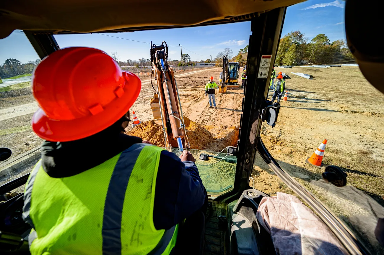 An operator is digging with a trencher/excavator. Three people wearing high-visibility vests are in front of the operator.