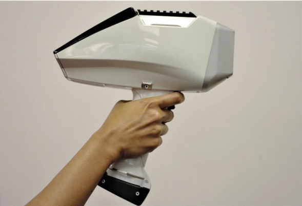 Digital Hand Spectrometer. A hand holds up a device with a hand grip similar to those on household drills. The digital hand spectrometer has a flat grey side that measures the metal and a screen which faces the operator.