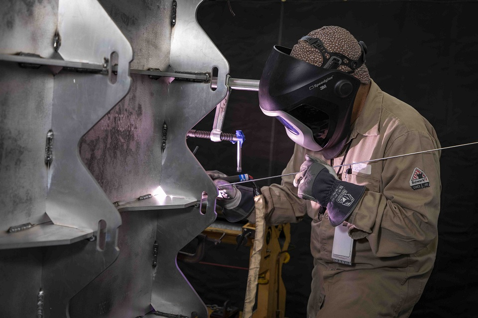 A welder wearing a face mask, leather gloves, a long-sleeved shirt, and a hair covering holds a torch in their right hand and an electrode in their right hand as they stand comfortably with their right-hand braced. They are in a shop setting and welding an upright aluminum weldment.