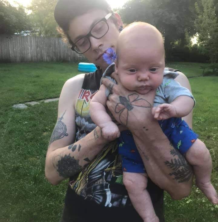 A white man holds an infant while standing in a green backyard.