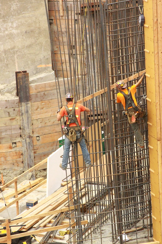 Two men working on a construction site wear fall protection harnesses to prevent injury.