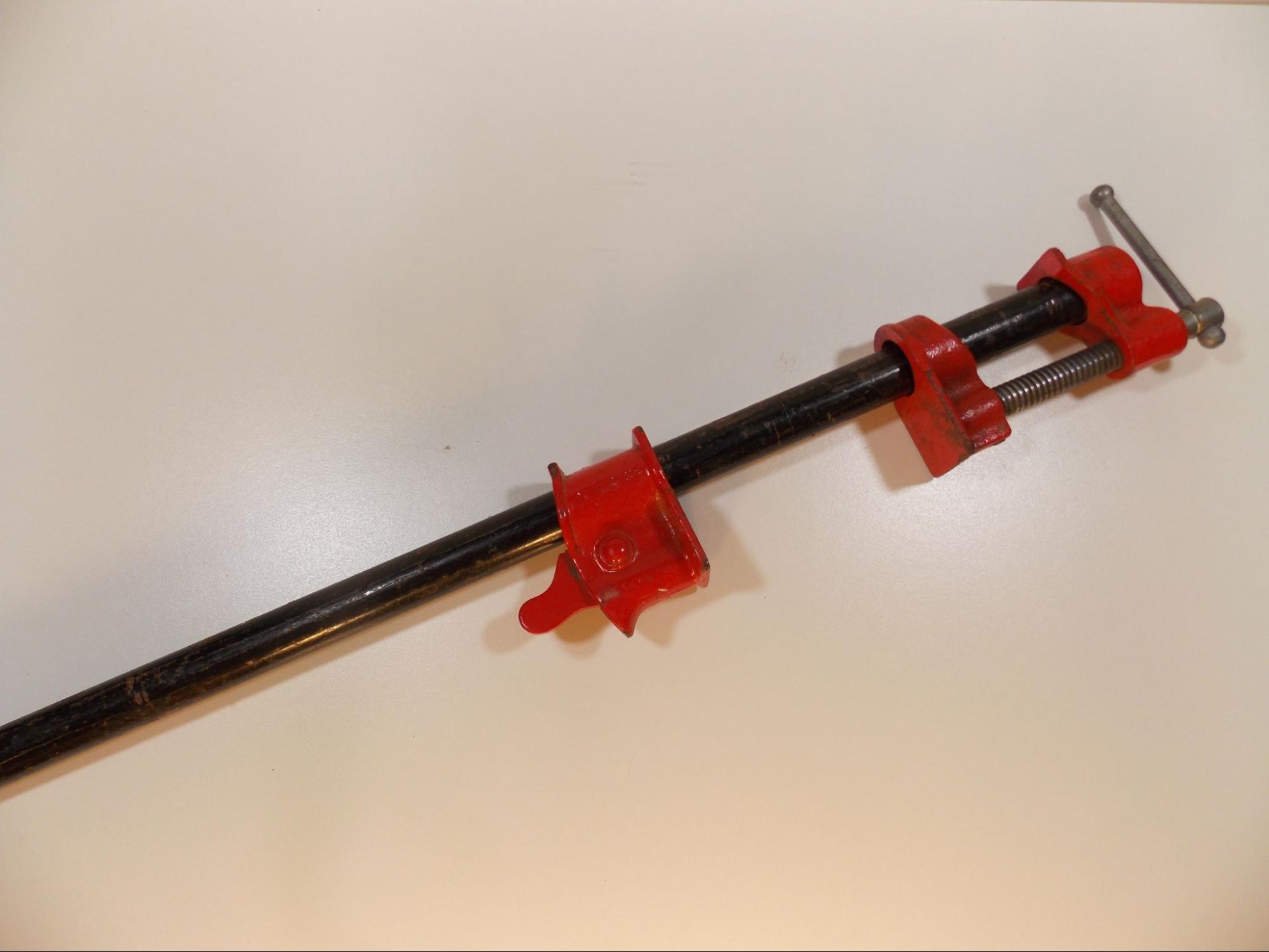 A common bar clamp. This clamp is a metal rod with two red metal jaws affixed to it. Each jaw has flat faces pointed toward each other. The lower jaw can slide up and down along the bar, while the upper jaw is mounted to the top of the bar and has a threaded screw to tighten or loosen the clamp.