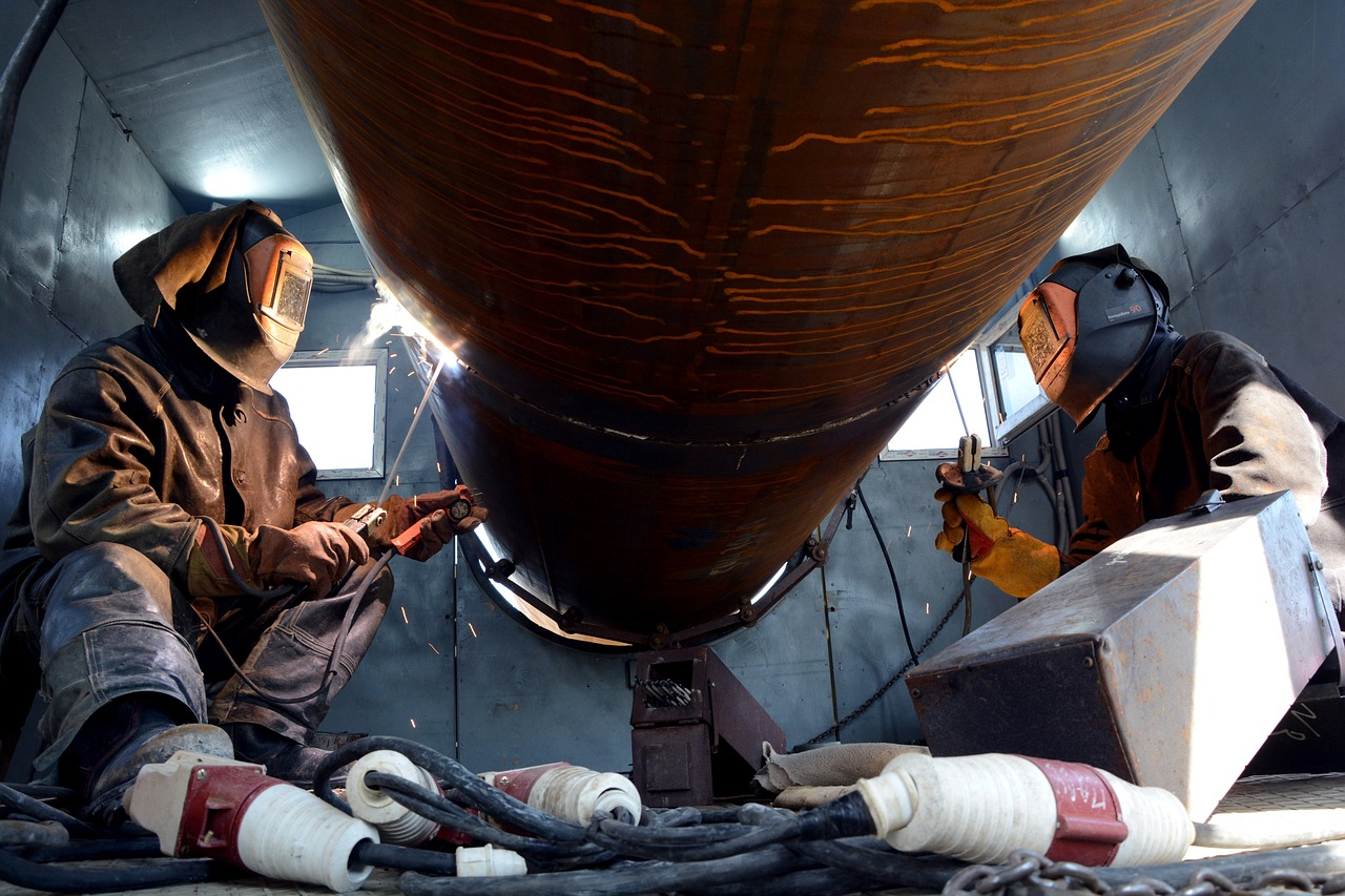 two pipeline welders using the SMAW process to splice piping together. The workers are wearing full PPE including welding helmets, gloves, protective pants, and leather welding aprons. They are sitting on the ground across from each other, on either side of the massive pipe. Both welders are in the process of welding, so their arcs can be seen as well as the sparks coming off of the metal pipe.