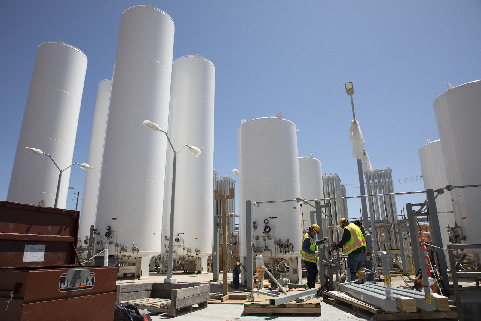 Two workers wearing yellow construction vests and hardhads work in a gas yard on gas yard piping. In the foreground, the workers are preparing some tubing. Surrounding them are eight white tube-shaped buildings.