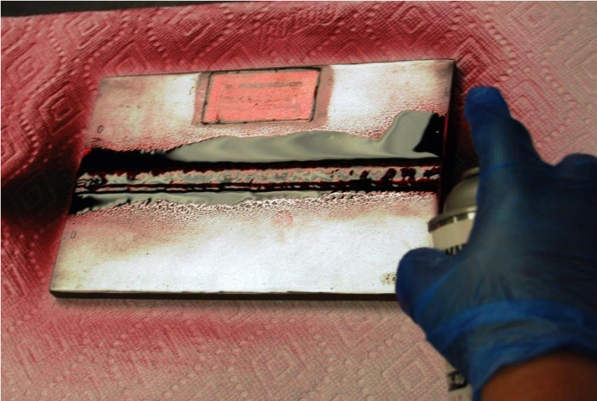 A hand wearing a blue latex-style glove uses an aerosol spray can to spray Red Liquid Penetrant Application on a flat weldment. The weldment is sitting on a paper towel.